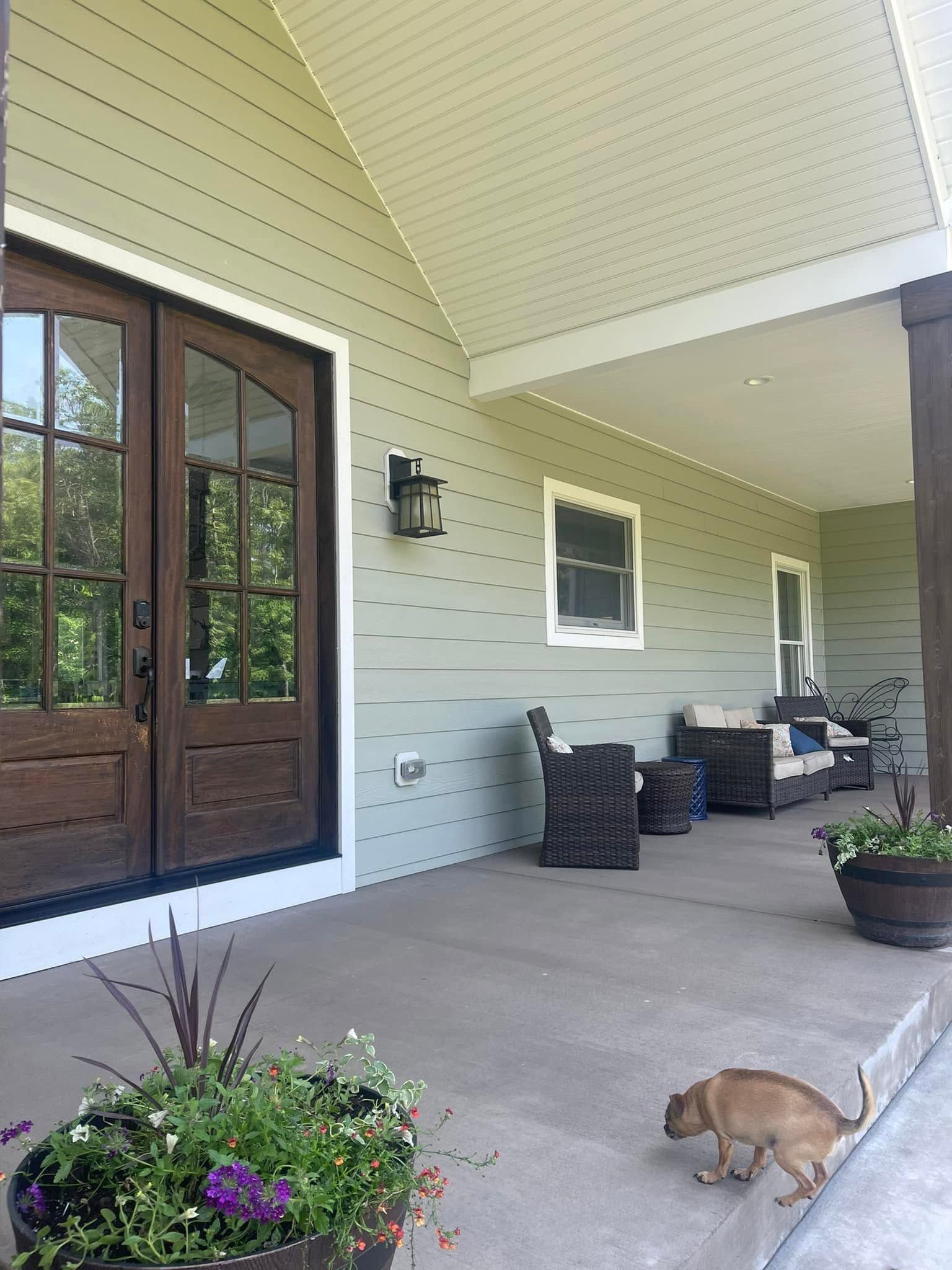Covered front porch with sage green lap siding, white trim, wood double doors, and a clean gray concrete floor