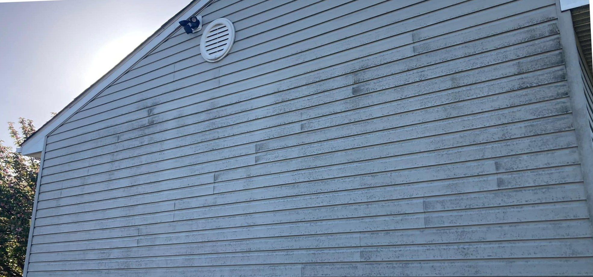 Gray vinyl gable with a round vent near the peak, speckled with dark mildew along most of the laps