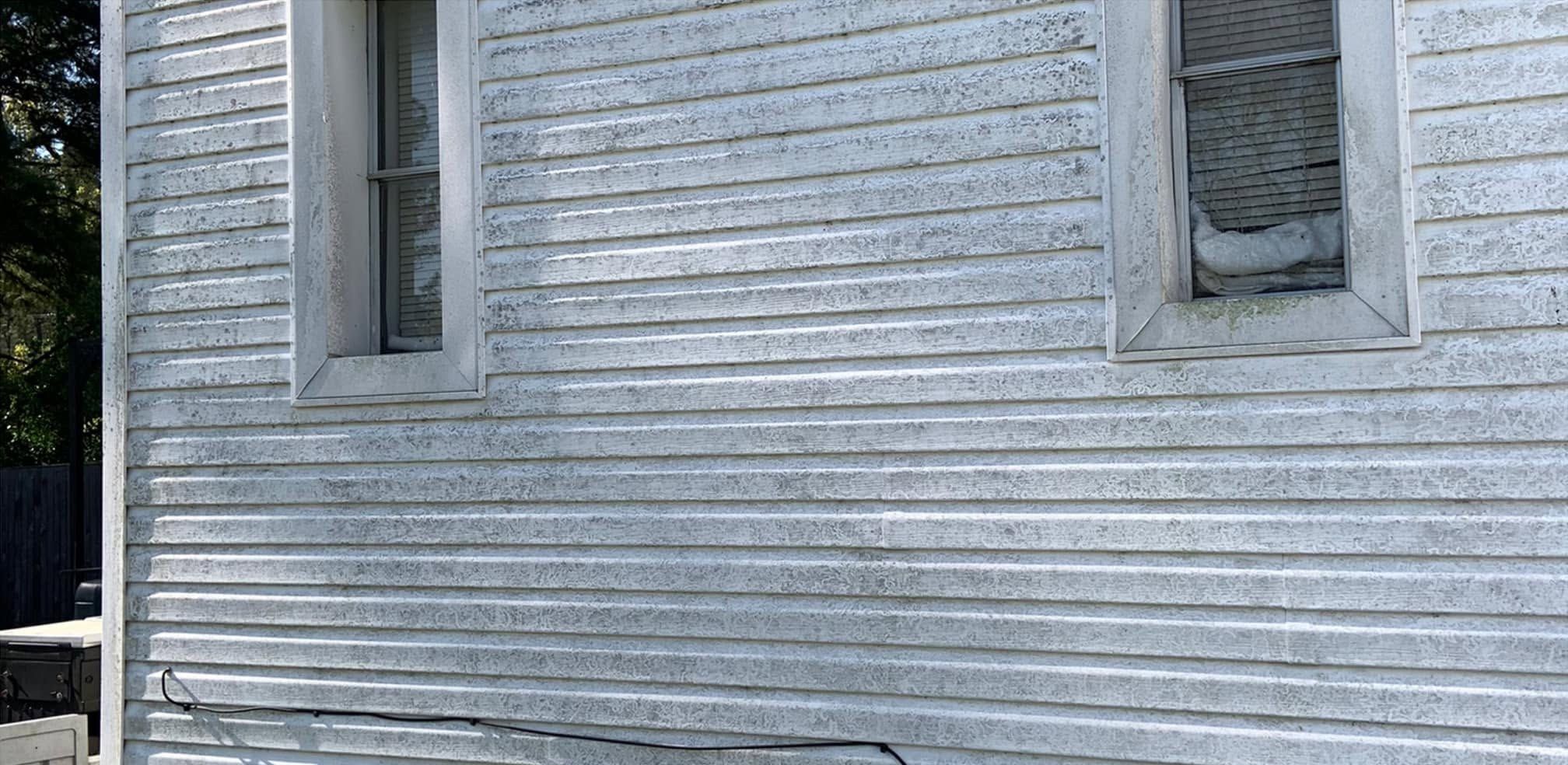 White vinyl siding between two windows, gray with mildew along every lap