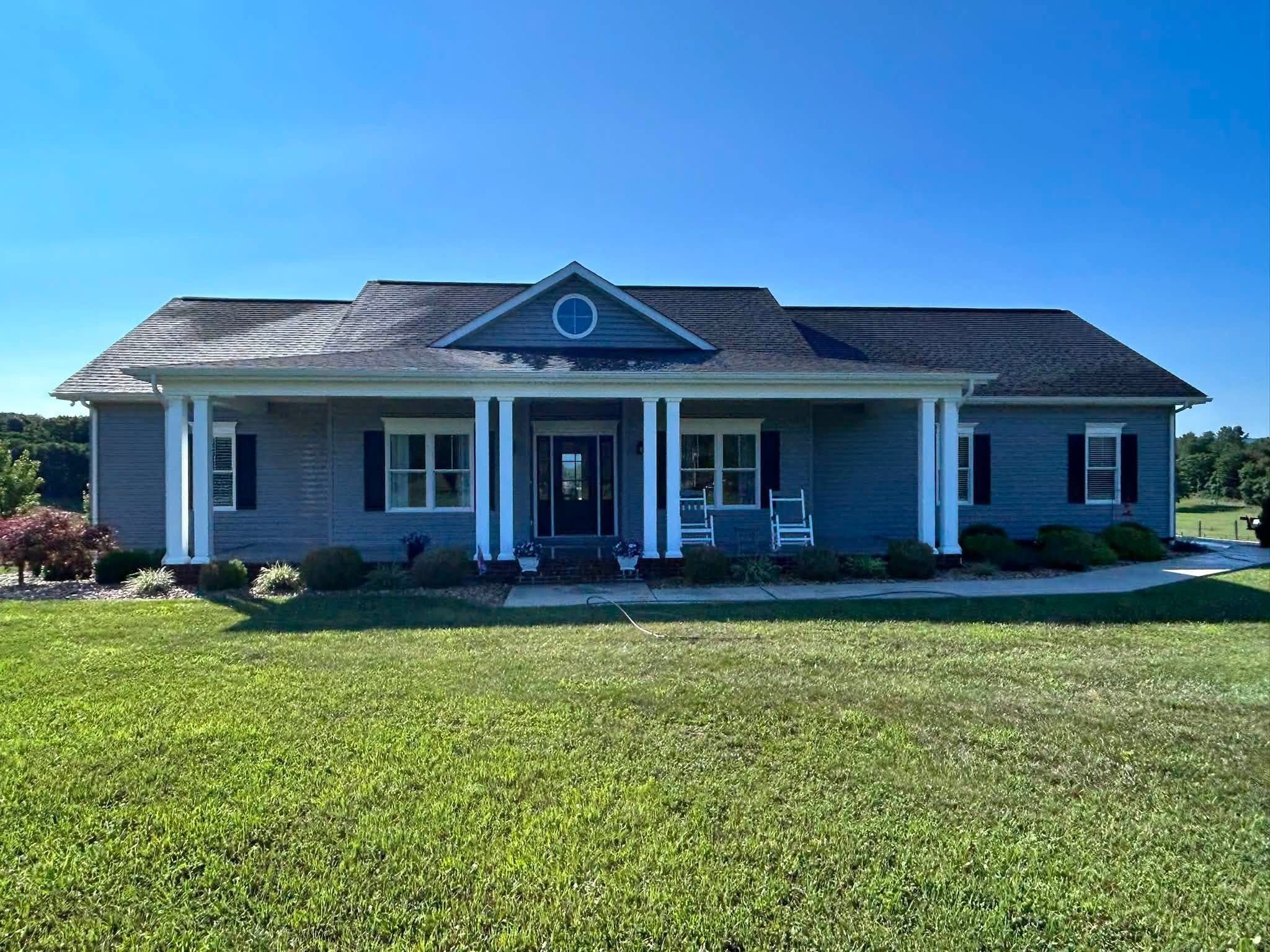 Single-story house with blue-gray siding and a full-width front porch with white columns and rocking chairs, set on a mowed lawn under a clear sky