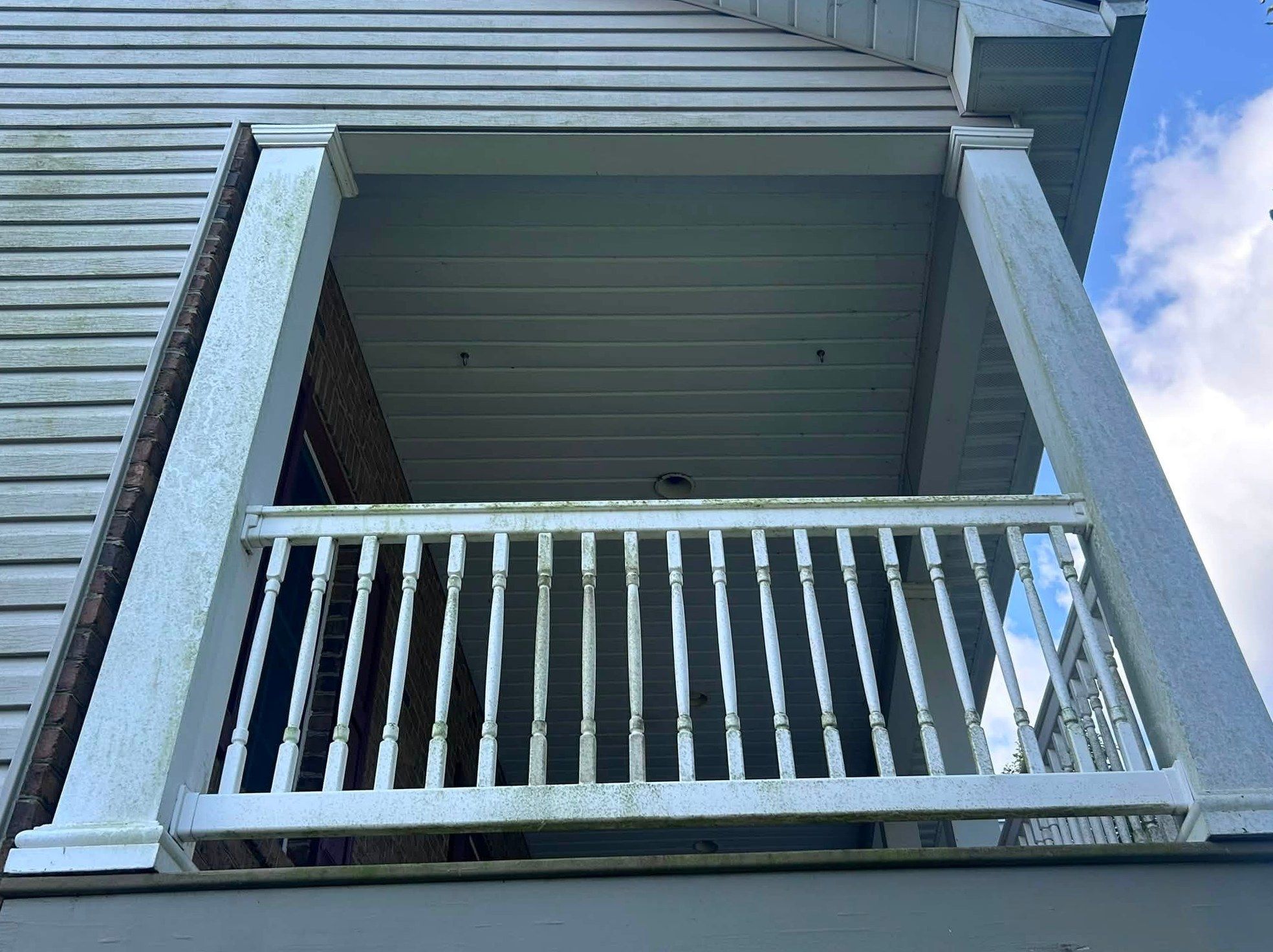 Covered balcony seen from below, white columns and railing streaked and spotted with green and black mildew against tan siding