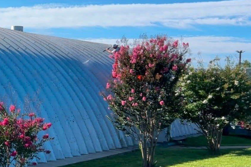 Crew member washing a curved metal roof beside flowering trees at the fairgrounds in Front Royal